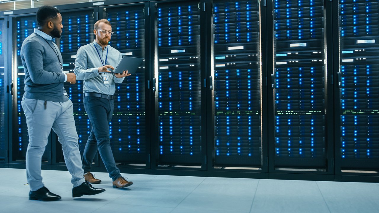  Two IT professionals walking through server room and consulting a laptop computer